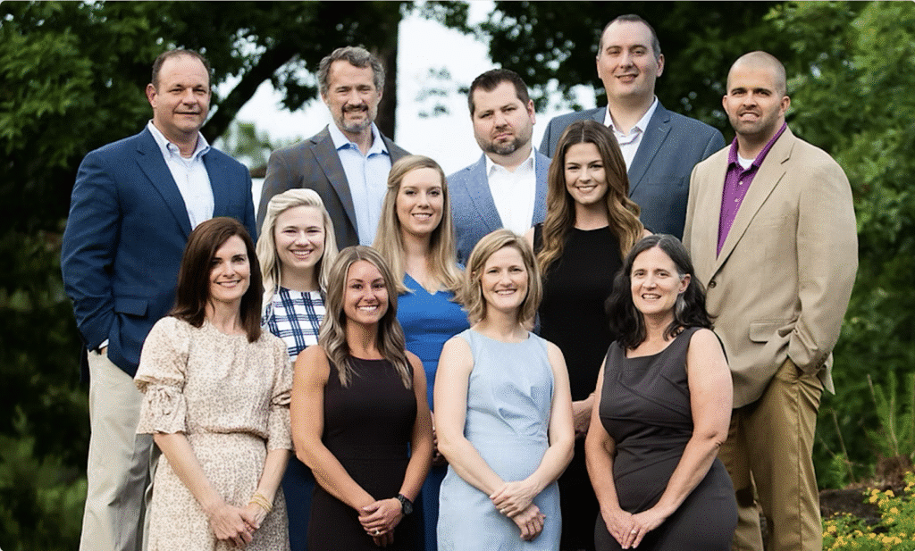Twelve adults, six men and six women, pose outdoors in semi-formal attire, smiling with trees and greenery behind them.